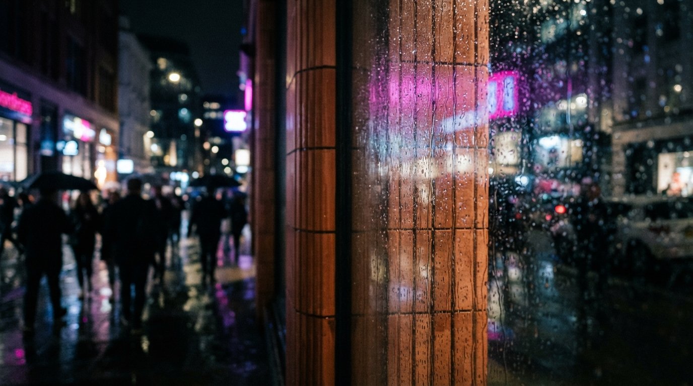 A close-up photograph of ribbed terracotta wall panels behind rain-speckled glass reflecting bright neon lights at night.