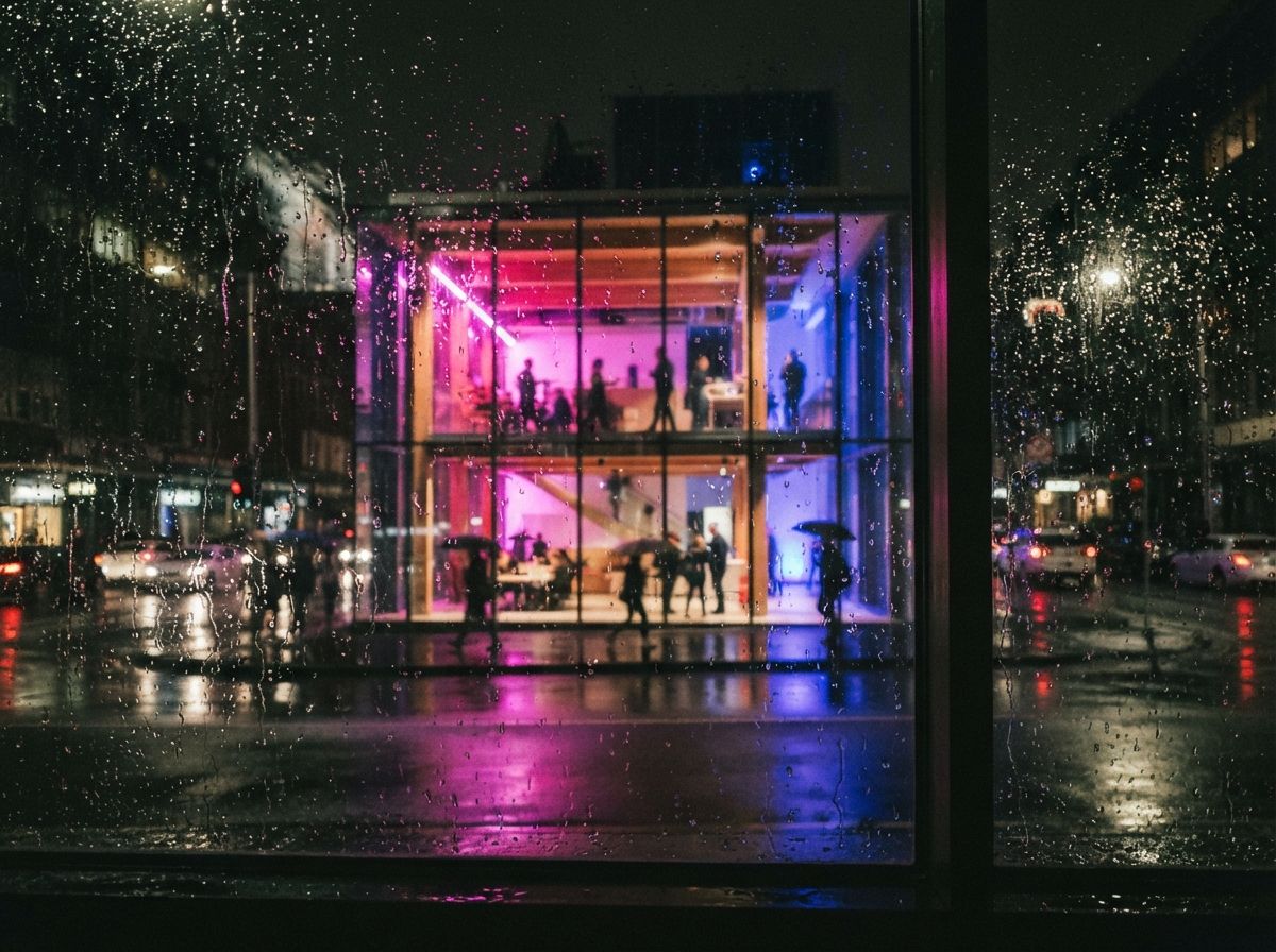 A night photograph shot through wet glass showing a neon-lit architectural studio interior with blurred silhouettes.